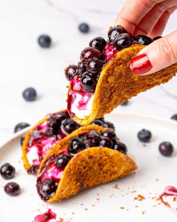Hand holding a taco filled with cheesecake and blueberries.