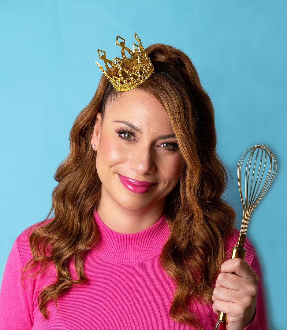 Emma holding a gold whisk in her left hand and wearing a small gold crown on her head.