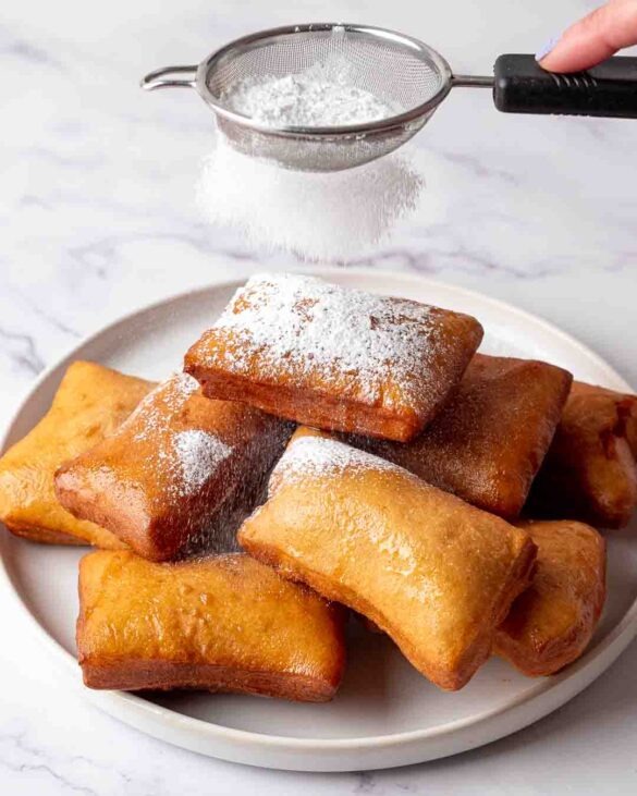 Icing sugar dusted over beignets piled on top of each other on a plate.