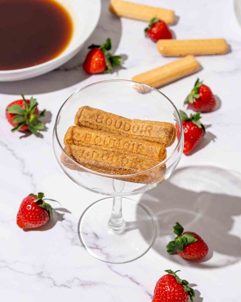 Three sponge fingers In a cocktail glass surrounded by strawberries on a marble counter.