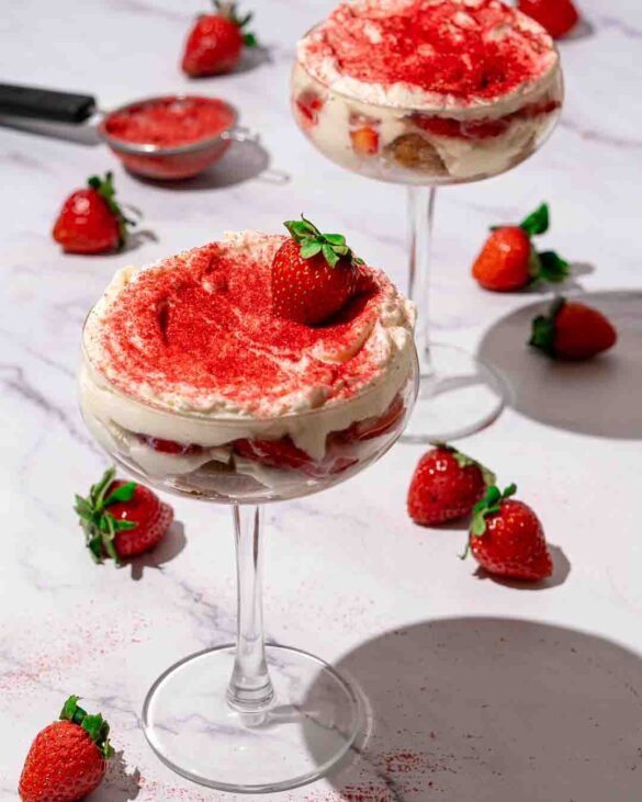 Strawberry tiramisu in cocktail glasses surrounded by strawberries on a white marble counter.