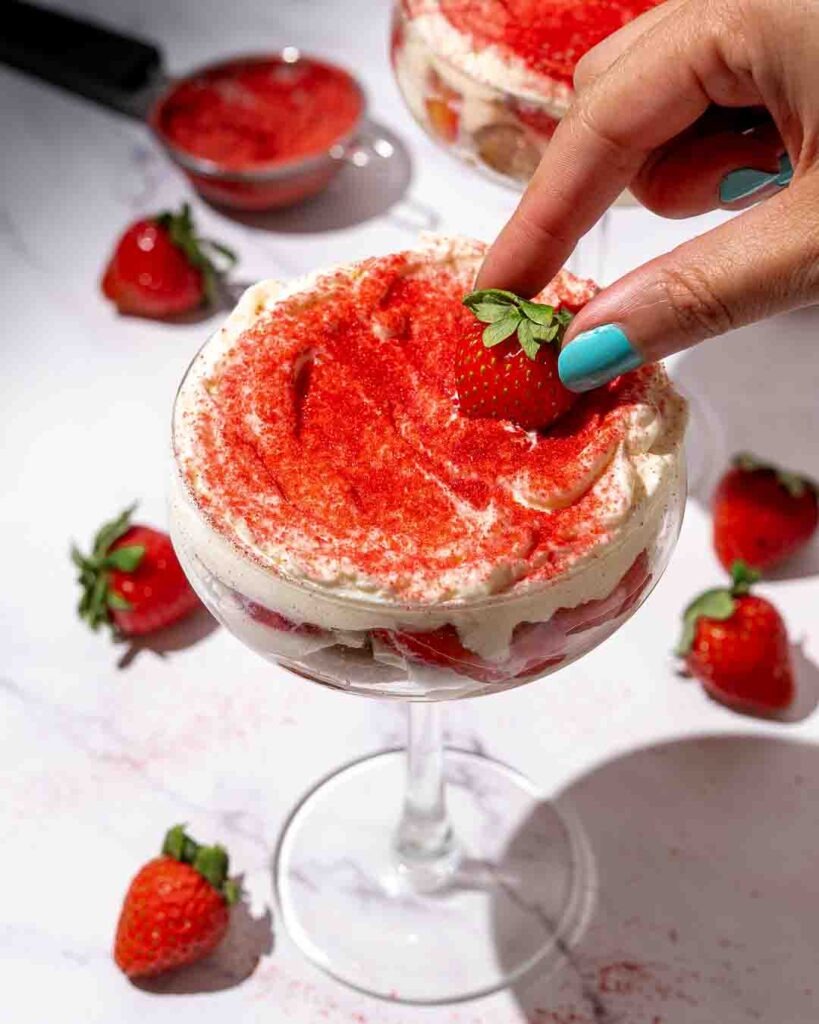 Strawberry tiramisu in cocktail glasses surrounded by strawberries on a white marble counter.