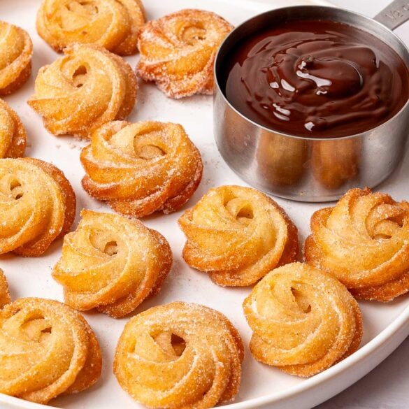 Mini churro swirls on a plate with a chocolate dip in a silver measuring cup.