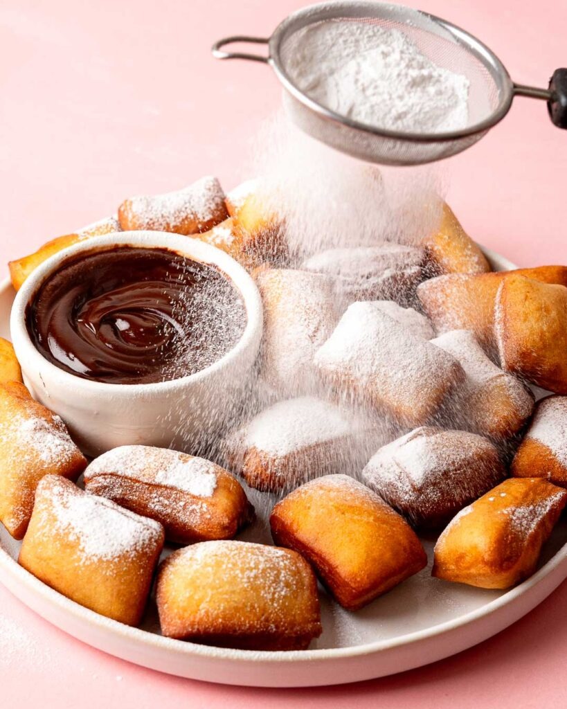 Mini Beignets piled up on a plate next to a chocolate ganache dip being dusted with icing sugar.