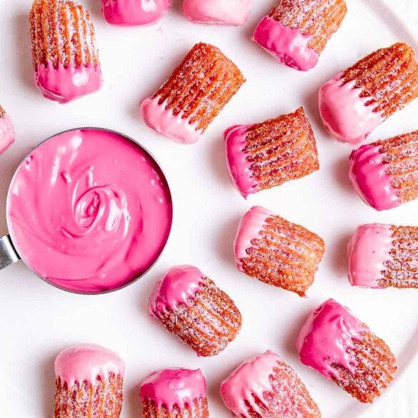 Mini pink churros dipped in pink chocolate on a plate surrounding a pink chocolate dip in a silver measuring cup.