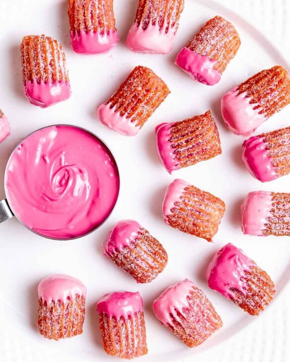 Mini pink churros dipped in pink chocolate on a plate surrounding a pink chocolate dip in a silver measuring cup.