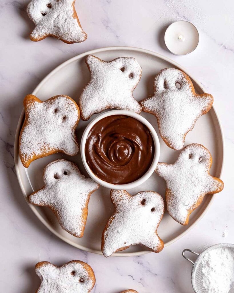 Ghost shape beignets on a plate with a chocolate dip in the middle.