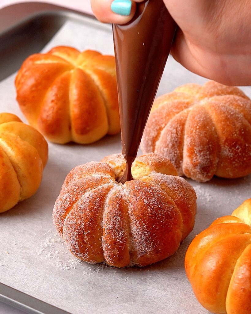 Pumpkin shaped buns being filled with Nutella using a piping bag.