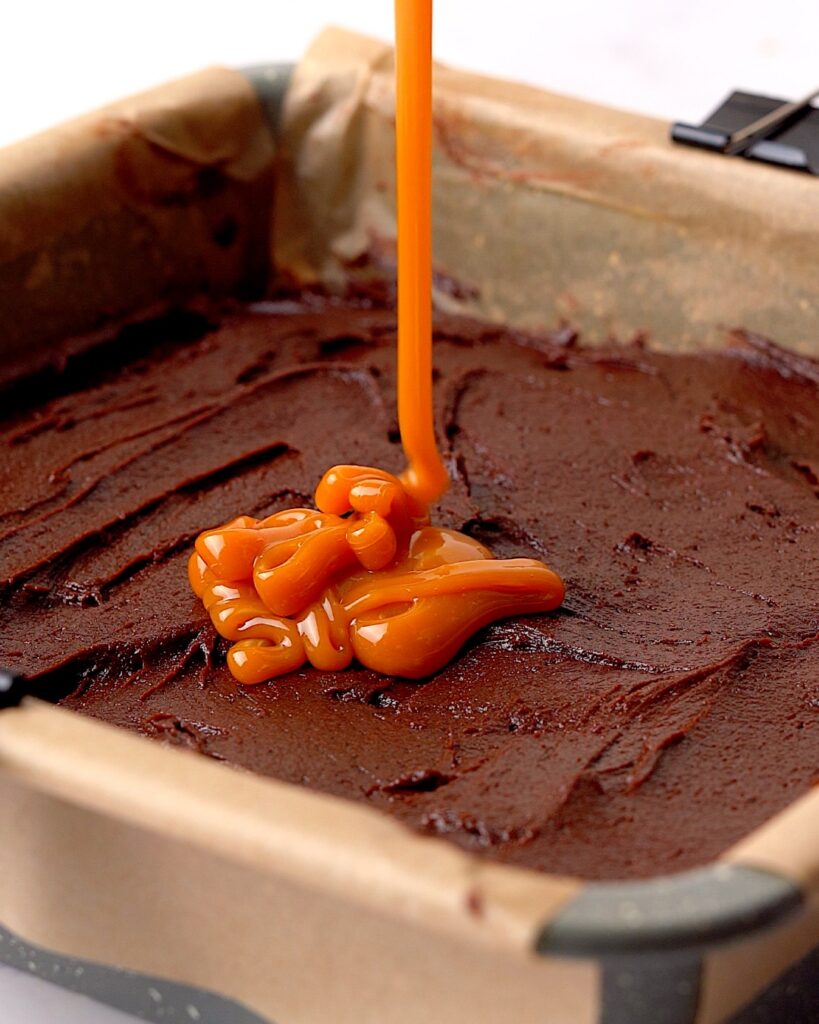 Caramel being poured on top of brownie batter in a baking tin.
