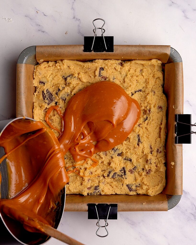 Caramel sauce being poured over cookie dough in a brownie tin.