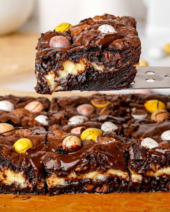 Mini Egg Cheesecake Brownies on a board, with one brownie being lifted above the rest using a spatula, showcasing the fudgy layers and colorful Mini Eggs.