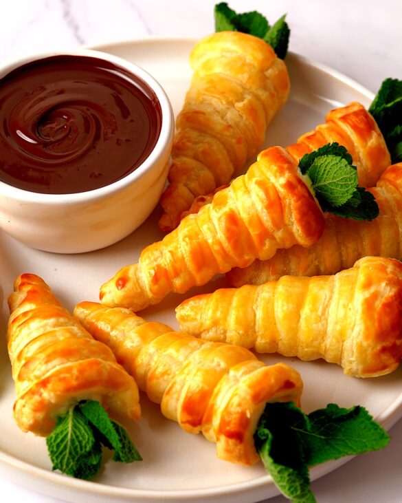 Puff pastry carrots on a plate with a pot of melted chocolate for dipping, perfect for an Easter treat.