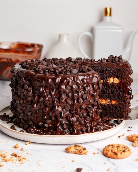 Chocolate chip cookie cake with a slice being pulled out, showing layered chocolate cake and chocolate chip cookies inside, topped with ganache and chocolate chips.