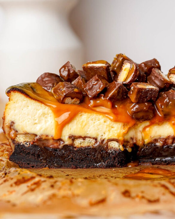Twix brownie Basque cheesecake cut in half, showing layers of fudgy brownie base, creamy cheesecake, chunks of Twix inside, caramel drizzle, and Twix pieces on top.