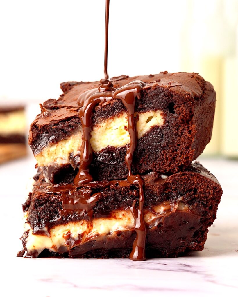 Cheesecake stuffed brownies stacked on top of each other with a drizzle of melted chocolate being poured on top.