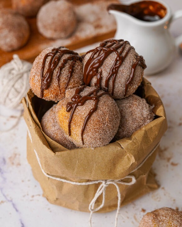 Air fryer doughnut holes covered in cinnamon sugar, drizzled in melted chocolate in a brown paper bag.