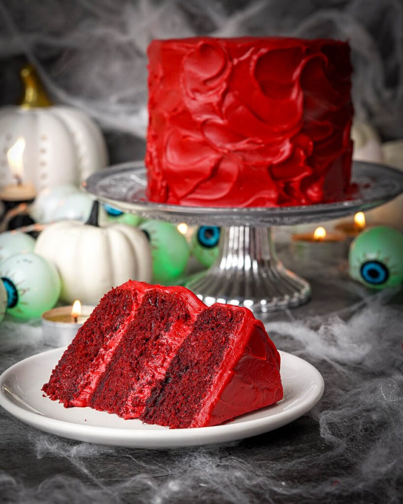 Mini red velvet cake with a messy red velvet cream cheese ganache on a silver cake stand and a slice of the cake on a white plate.