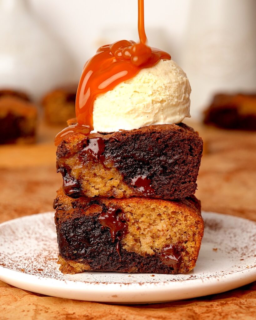 Sticky toffee pudding brookies stacked on a plate with ice cream on top and a drizzle of caramel sauce being poured on top.