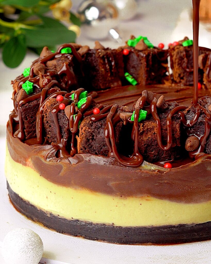 Whole no-bake pistachio cheesecake with a brownie base, topped with glossy chocolate ganache, brownie pieces and red and green Christmas sprinkles in a wreath shape.