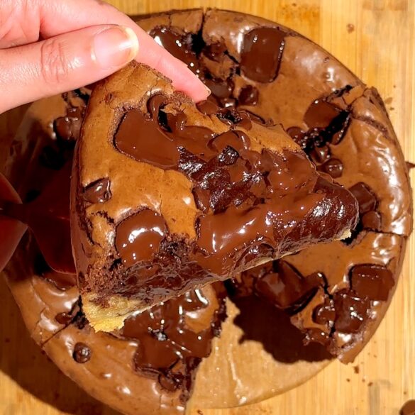 Hand lifting a thick chocolate chip cookie with gooey melted chocolate chunks on a wooden surface.