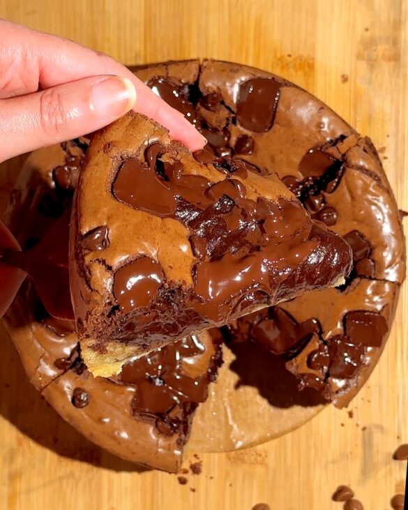 Hand lifting a thick chocolate chip cookie with gooey melted chocolate chunks on a wooden surface.